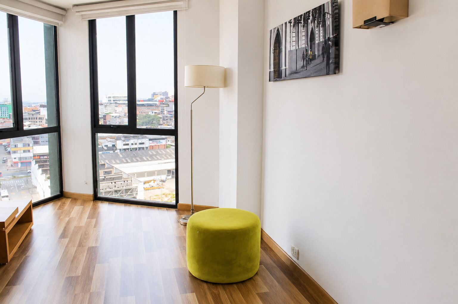 A corner room with large windows, a floor lamp, a green ottoman, and a framed picture on the wall.
