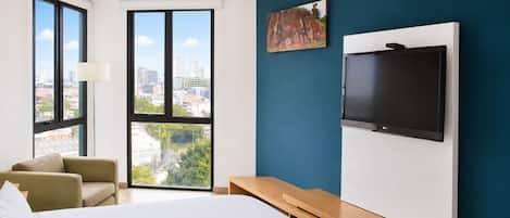 Hotelkamer met een bed, bureau, stoel, televisie en groot raam met uitzicht op de stad.