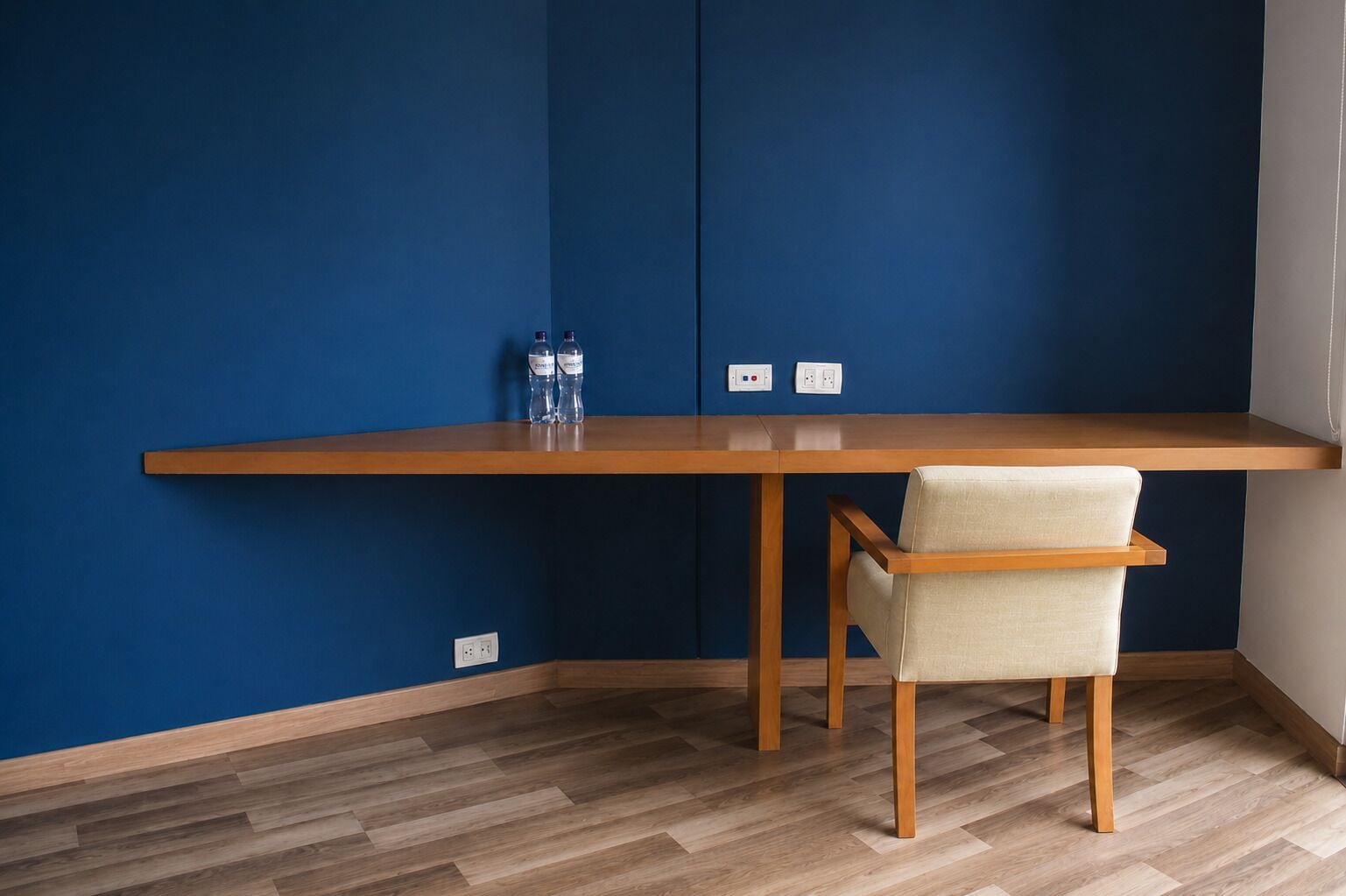 Una habitación con un escritorio de madera, una silla, dos botellas de agua y enchufes en la pared.