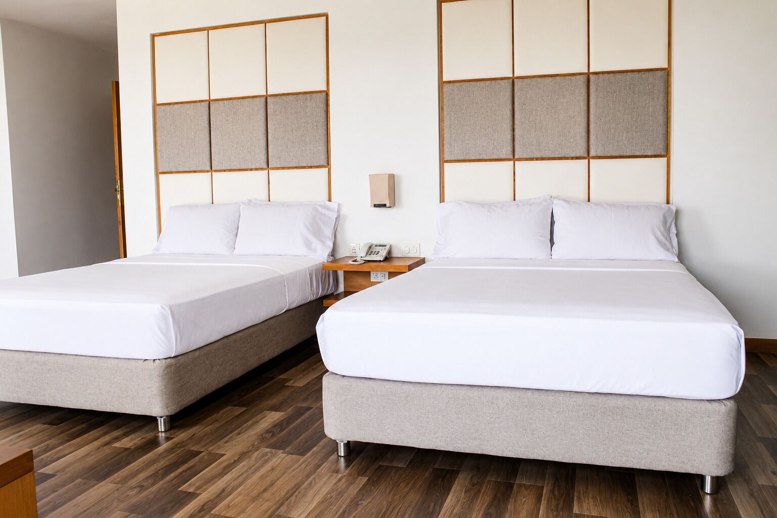 Two twin beds with grey bedding, a wooden floor, a bedside table with a phone, and a wall panel.