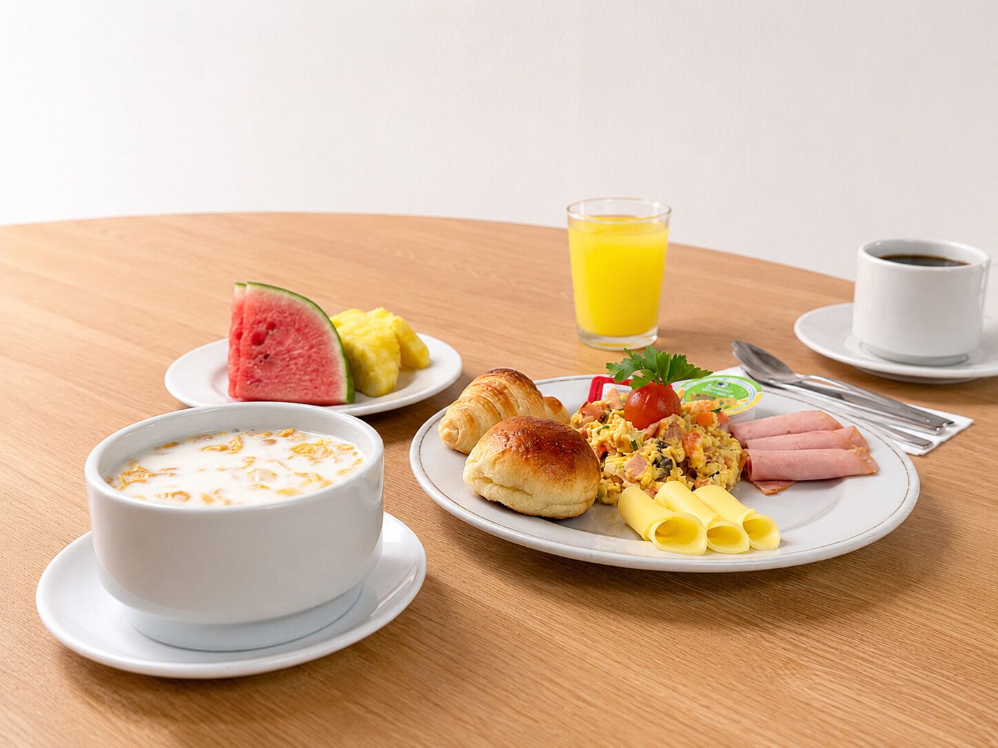 Un desayuno con un plato de huevos, jamón y verduras, un vaso de jugo de naranja, un tazón de yogur y una rebanada de sandía.