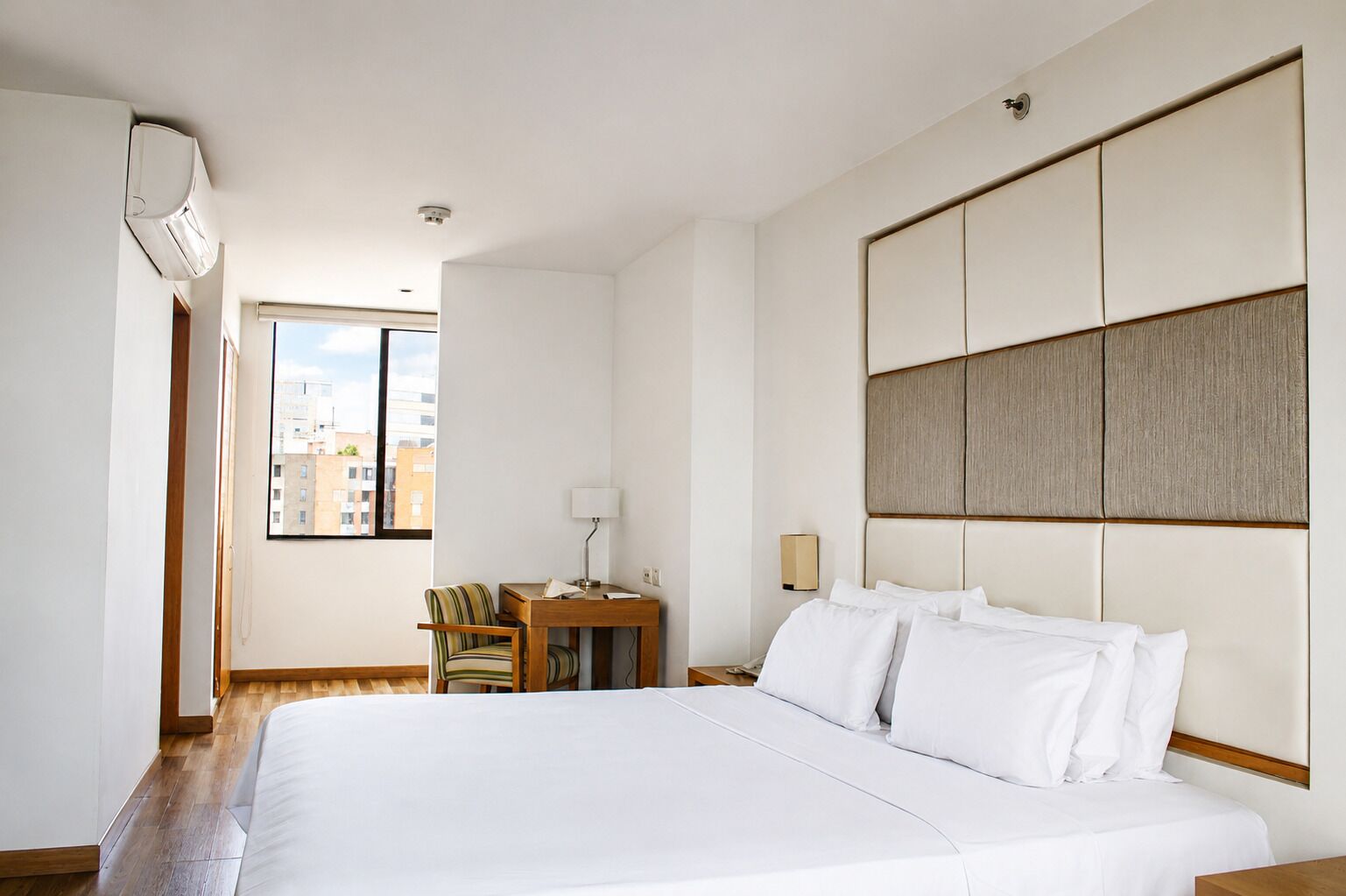 Quarto de hotel com uma cama grande, uma mesa com cadeira, uma luminária e vista para prédios pela janela.