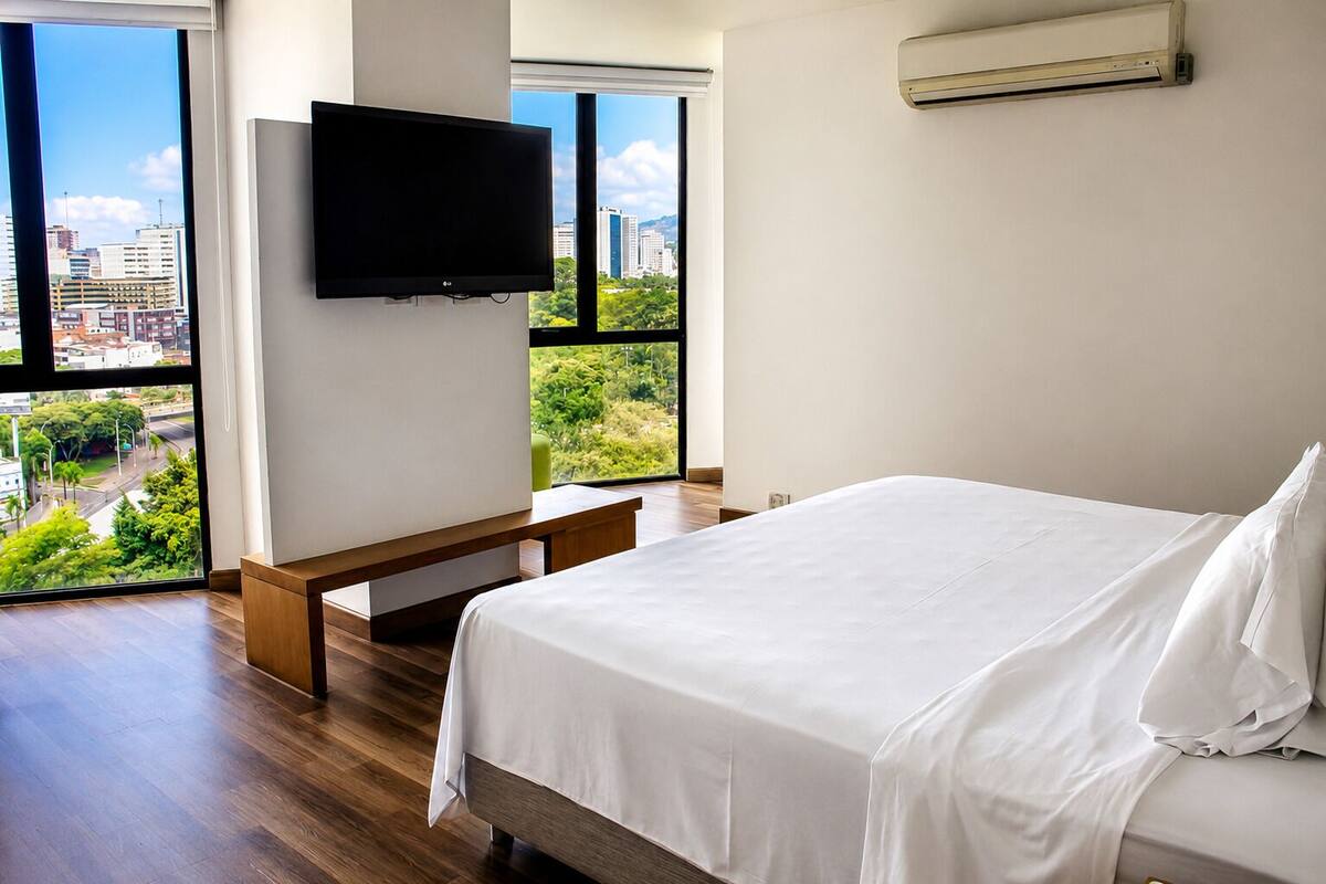 Habitación de hotel con cama, televisor montado en la pared y ventanas amplias con vista a la ciudad.