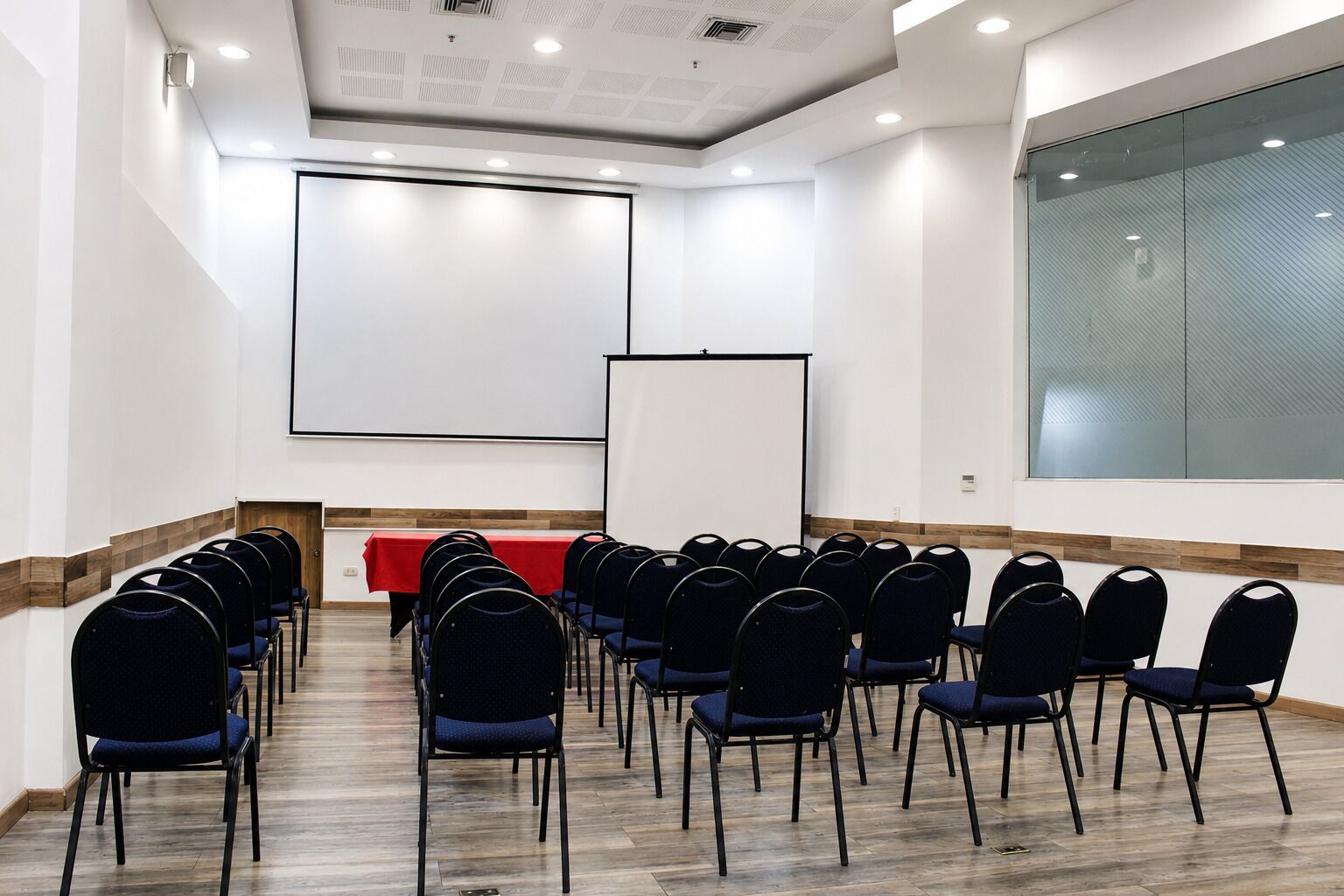 A conference room with rows of chairs, a red table, and two projection screens.