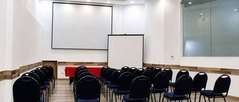 Un salón de conferencias con filas de sillas, una mesa roja y dos pantallas de proyección.