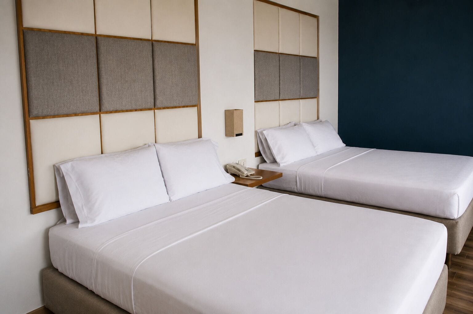 A hotel room with two beds, a bedside table with a phone, and a wall-mounted panel.