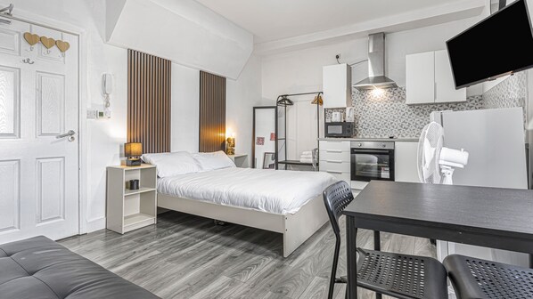 A bedroom with a bed, bedside tables, a desk, a chair, a TV, and a kitchenette with a microwave and oven.