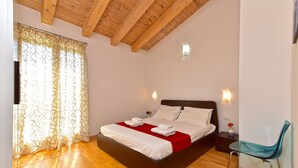 Ein Schlafzimmer mit Holzbalkendecke, einem Bett mit weißen Leinen, einer roten Tagesdecke, einem Glastisch mit Stuhl und einer Wandleuchte.