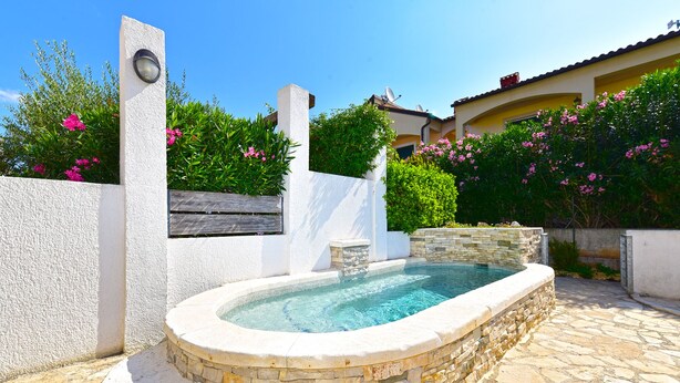 Una piscina de piedra con un muro de piedra y un banco de madera, rodeada de vegetación y un edificio de fachada amarilla.