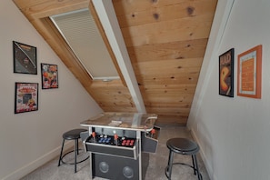 A room with a sloped ceiling, wooden beams, a foosball table, two stools, and wall-mounted posters. - Mountain Retreat, Theater Room, Hot Tub, Pac-Man Arcade & Stone Fireplace: Sugar Pine Lodge (South Lake Tahoe)