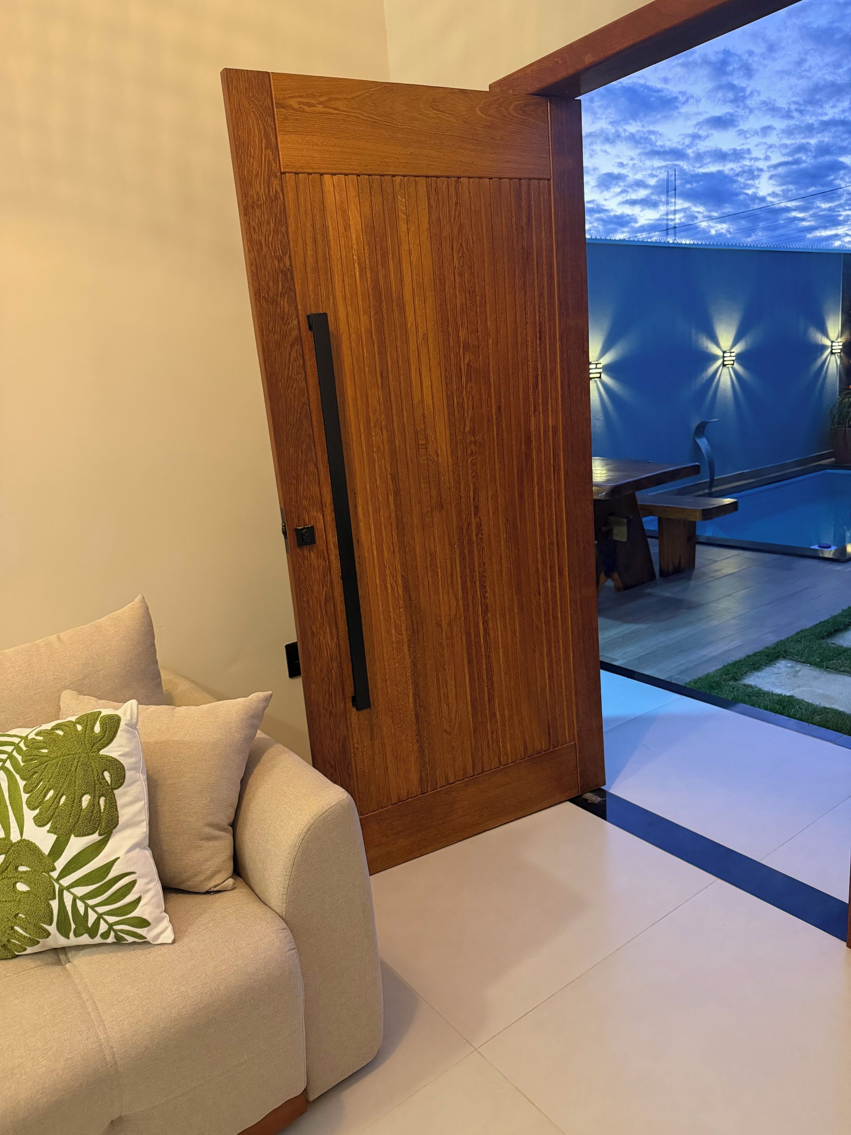 A modern interior with a wooden door, a beige sofa with a green leaf pillow, and a pool area with lights.