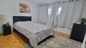 Ein Schlafzimmer mit Bett, Nachttisch, Lampe, einem Fenster mit Vorhängen und einem Poster an der Wand.