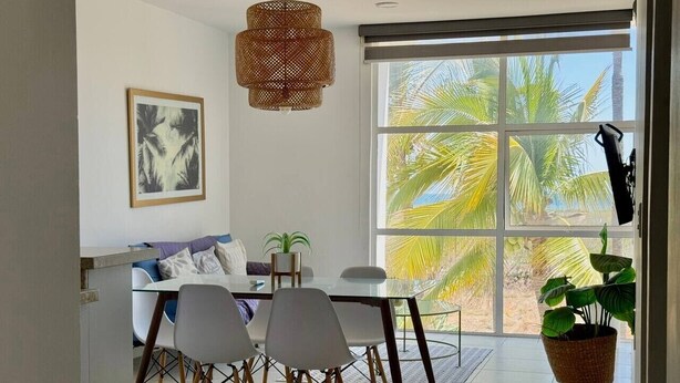 A modern dining area with a glass table, white chairs, a woven pendant light, and a large window with a view of palm trees. - Tu Escape Perfecto en Mazatlán by Partner Host (Mazatlán)