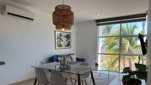 A modern living room with a glass dining table, white chairs, a woven pendant light, a sofa with cushions, a framed picture on the wall, and a large window with a view of palm trees. - Tu Escape Perfecto en Mazatlán by Partner Host (Mazatlán)