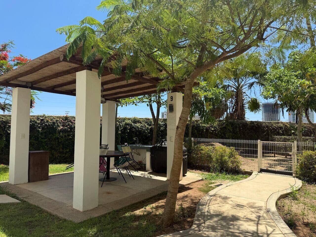 Un espace extérieur abrité, doté d’un barbecue et de sièges, entouré de verdure et bordé d’un sentier.