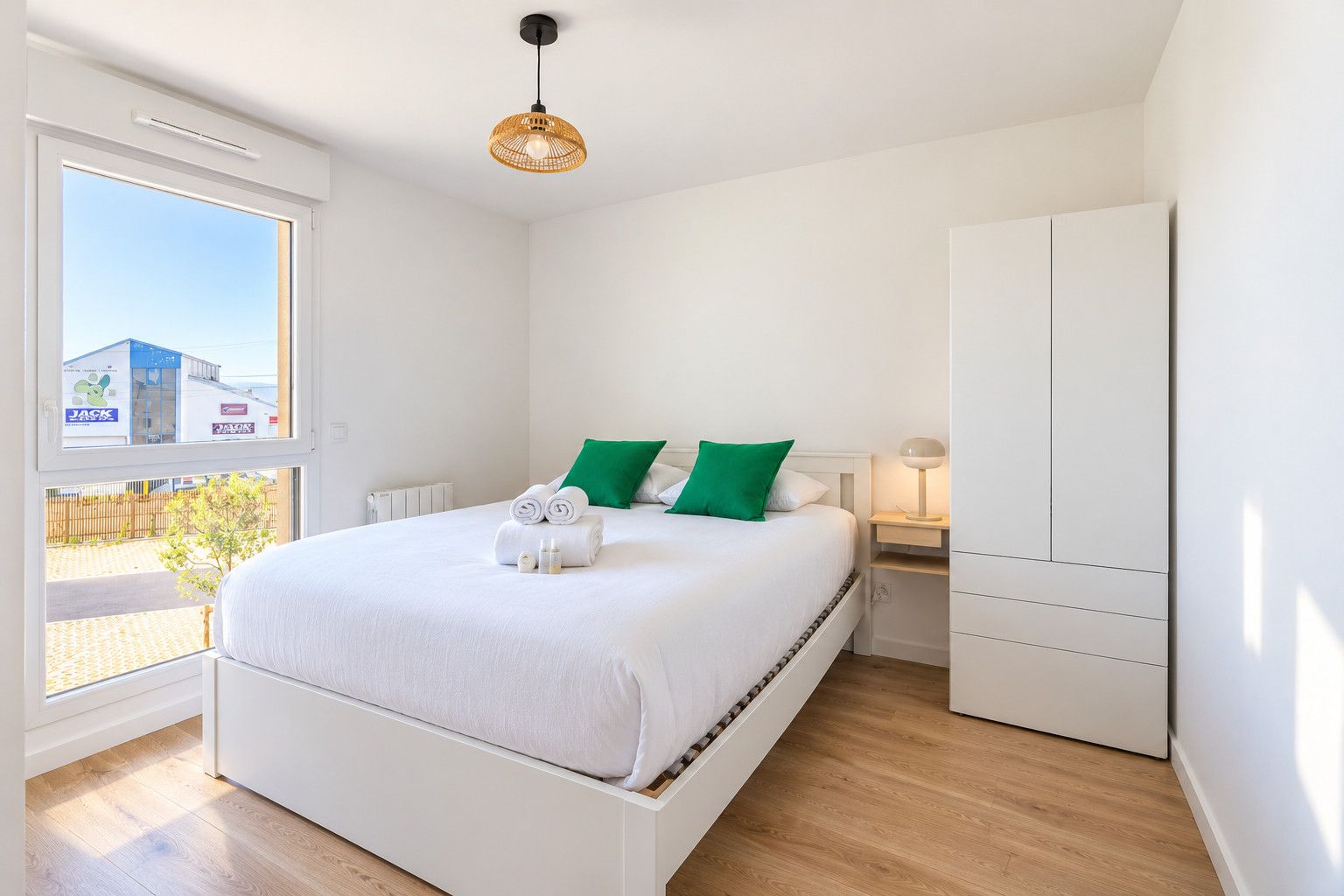 A bedroom with a bed, a wardrobe, a bedside table with a lamp, and a window with a view of buildings.