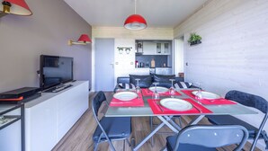 Un coin repas avec une table dressée pour quatre personnes, un téléviseur posé sur un meuble, et une cuisine en arrière-plan.