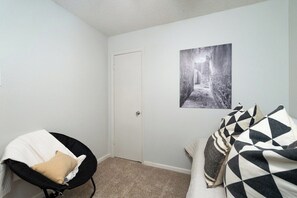 Ein Zimmer mit einer Tür, einem schwarzen Stuhl mit beiger Sitzkissen, einer gemusterten Tagesdecke und einem Schwarz-Weiß-Foto an der Wand.