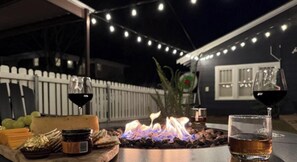 Un espacio al aire libre para comer con un hogar, una mesa con comida y bebidas, y luces colgantes.
