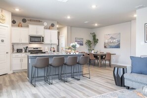 A modern kitchen with white cabinets, a central island with bar stools, a dining area with a table and chairs, and a living space with a sofa and a coffee table.