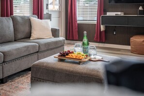 A living room with a grey sofa, a wooden side table with a fruit platter, a bottle, and glasses. - 5-Bedroom Themed Retreat - The Providence (Abilene)