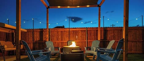 An outdoor patio with a fire pit, two Adirondack chairs, and string lights.