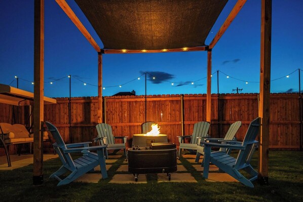 An outdoor patio with a fire pit, two Adirondack chairs, and string lights.