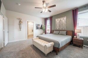 A bedroom with a bed, a bench, a ceiling fan, a nightstand, and a wall-mounted macrame piece. - 5-Bedroom Themed Retreat - The Providence (Abilene)