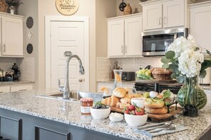 A kitchen with white cabinets, a granite countertop, a stainless steel sink, and a breakfast spread including bread, fruits, and a vase with flowers.