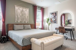 Quarto com cama, escrivaninha, cadeira, tapeçaria de macramé e uma obra de arte emoldurada.