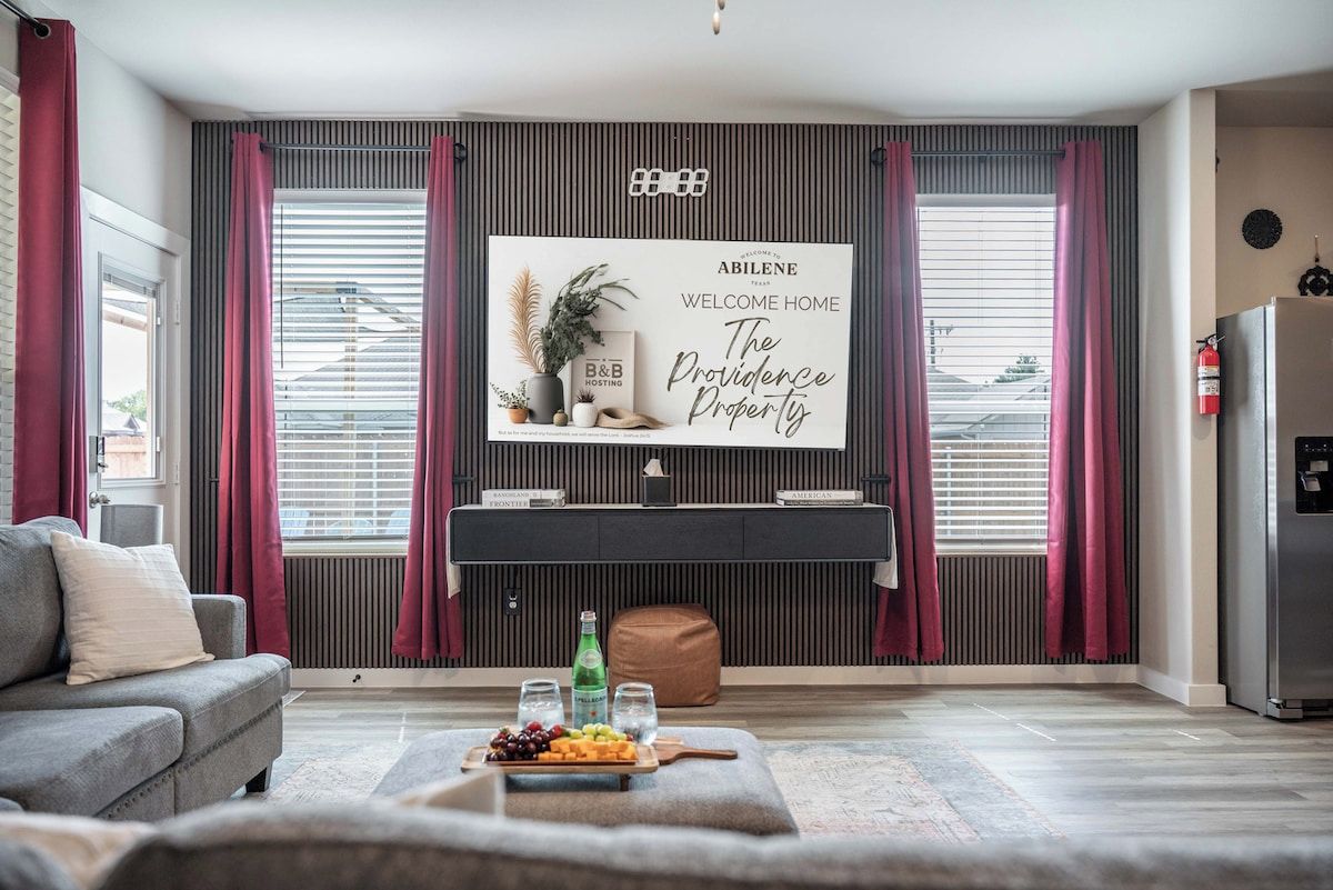 A modern living room with a flat-screen TV, a grey sofa, a wooden coffee table with snacks, and a large window with blinds.