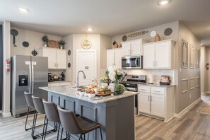 A modern kitchen with a central island, bar stools, a stainless steel refrigerator, and white cabinetry. - 5-Bedroom Themed Retreat - The Providence (Abilene)