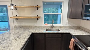 A kitchen with granite countertops, dark cabinetry, a stainless steel sink, wooden shelves, and a window with a view. - Charming 3-bedroom house in enjoyable Gig Harbor with AC (Gig Harbor)