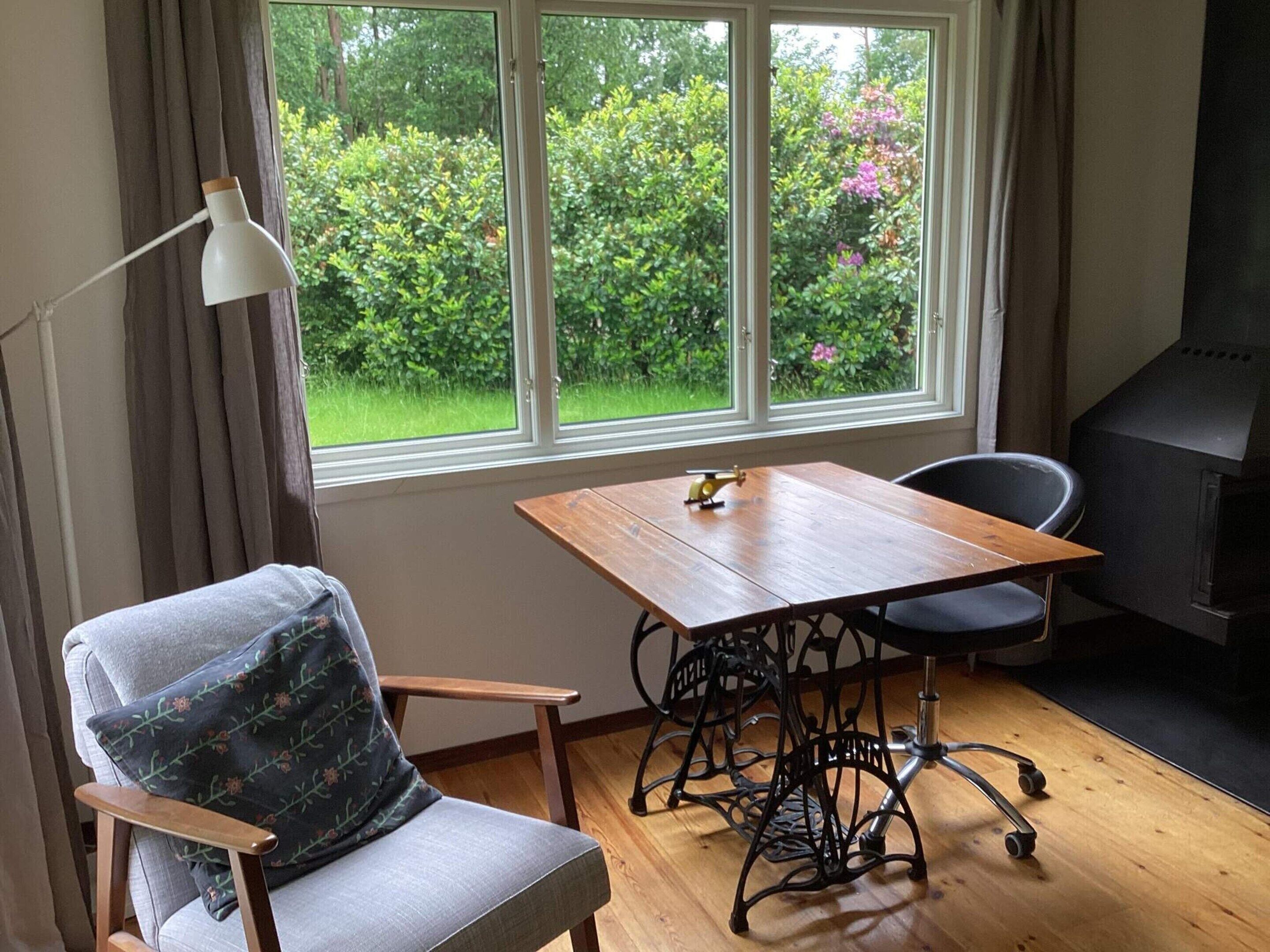 A room with a table, a chair, a lamp, and a view of greenery outside.