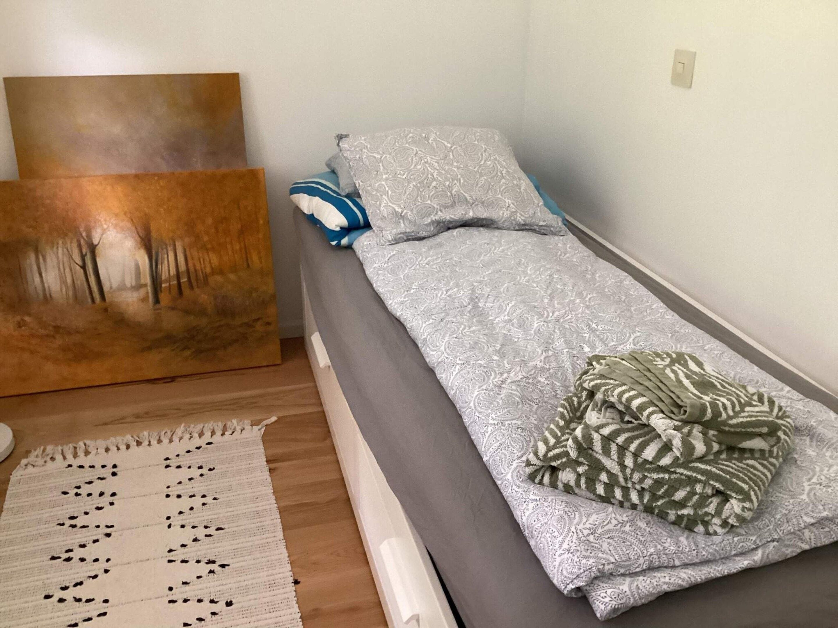 Una cama individual con colchón gris y ropa de cama estampada, una pared blanca y un cuadro a la izquierda.