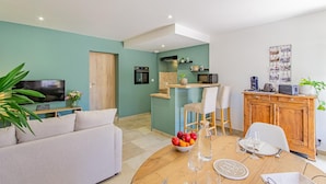 Een moderne woonkamer met een bank, een eettafel gedekt voor twee personen, een keuken met een magnetron en oven, en een televisie op een meubel.