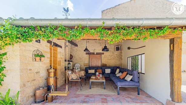 Een terras buiten met stenen muren, een houten bank, een bankstel en hangende planten.