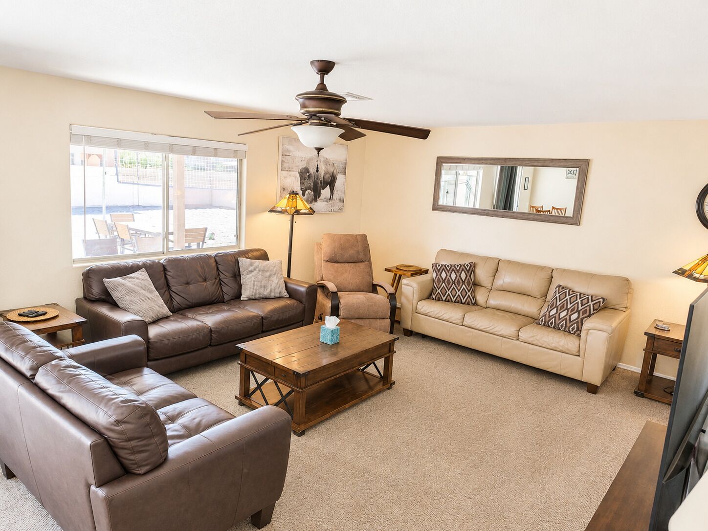 A living room with a brown leather sofa, a beige sofa, a wooden coffee table, a ceiling fan, and a large window with a view of outdoor seating.
