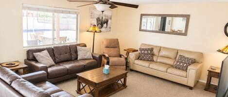 A living room with a brown leather sofa, a beige sofa, a wooden coffee table, a ceiling fan, and a large window with a view of outdoor seating.