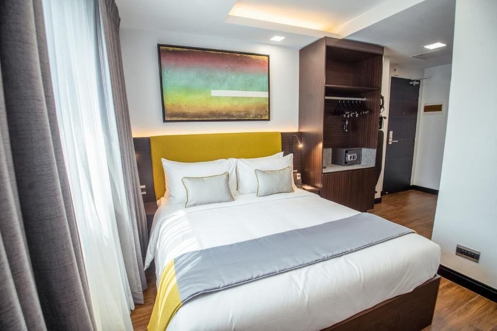 A hotel room with a large bed, a headboard, a painting, a wardrobe, and a kitchenette.