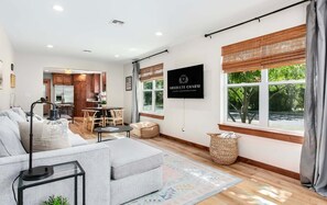 Una sala de estar moderna con un sofá, una mesa de centro y un televisor montado en la pared. Hay una zona de comedor con una mesa y sillas, y una cocina al fondo.