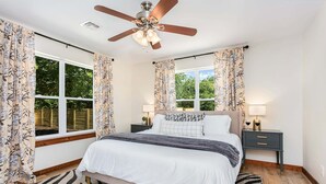 Ein Schlafzimmer mit einem Bett, Deckenventilator, zwei Nachttischen mit Lampen und einem Fenster mit Vorhängen.