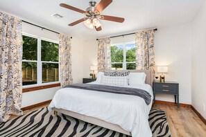 Ein Schlafzimmer mit einem Bett, Deckenventilator, zwei Nachttischen mit Lampen und einem Fenster mit Vorhängen.
