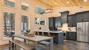 A modern kitchen with dark cabinets, stainless steel appliances, and a wooden dining table with benches. - NEW! 4 King Suites, Hot Tub, Fire Pit, Outdoor Lounge, Spacious (Broken Bow)
