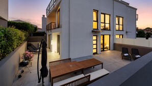A modern two-story building with a patio area featuring a wooden table and chairs, a barbecue grill, and a small garden with pink flowers. - House Explorer One (Istria - Funtana) (Funtana)
