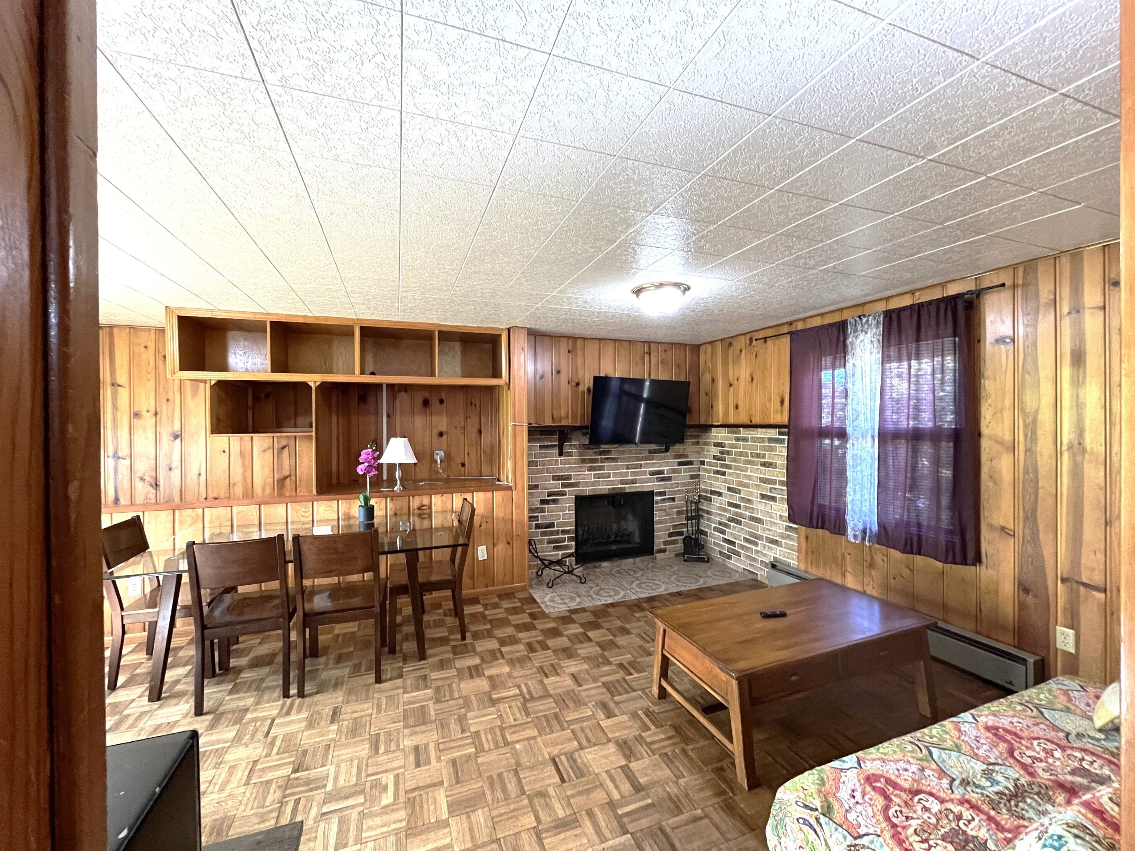 L'intérieur chaleureux d'un chalet avec un plancher en bois, une cheminée, un téléviseur fixé au mur et un coin repas comprenant des chaises et une table.
