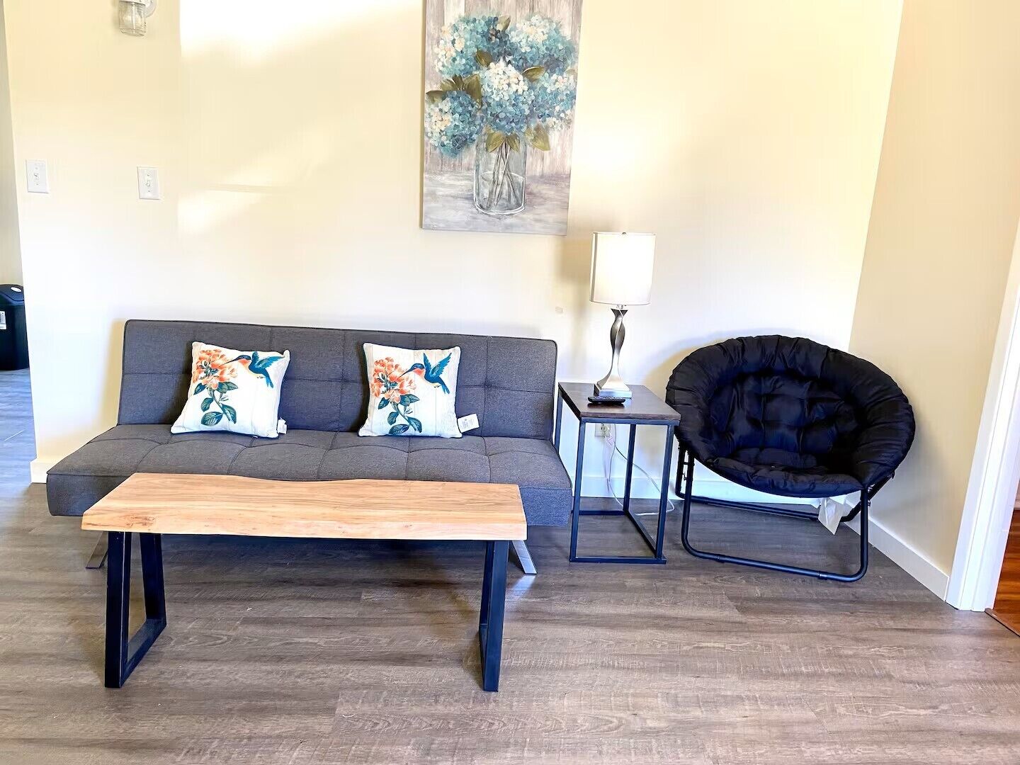 Un salon avec un canapé gris, deux coussins à motifs floraux, une table basse en bois, un fauteuil noir et une petite table d’appoint avec une lampe.
