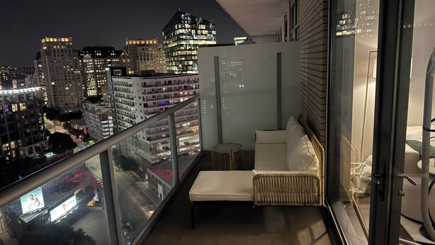 A balcony with a view of a cityscape at night, a cushioned seating area, and a glass railing.
