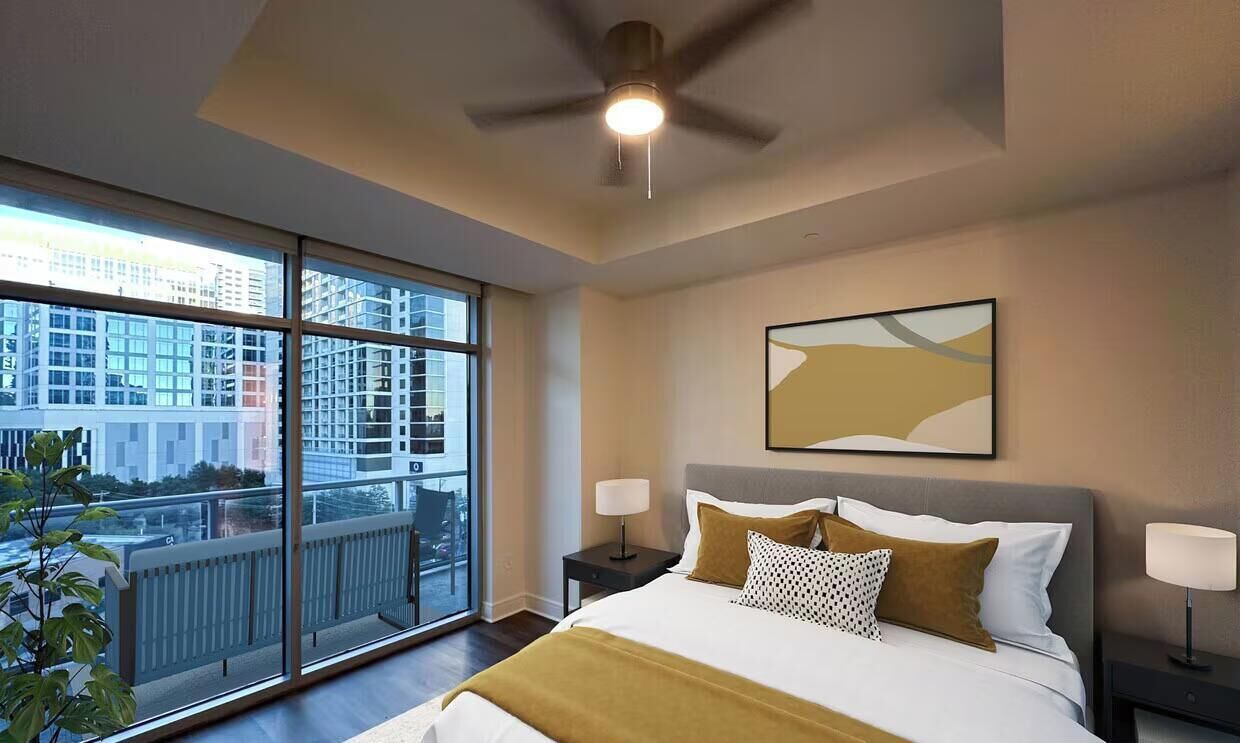 A hotel room with a large bed, two bedside tables, a ceiling fan, and a view of the city through floor-to-ceiling windows.