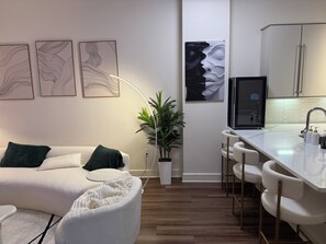 A modern living space with a sofa, a white armchair, a plant, and a kitchen area with a bar.